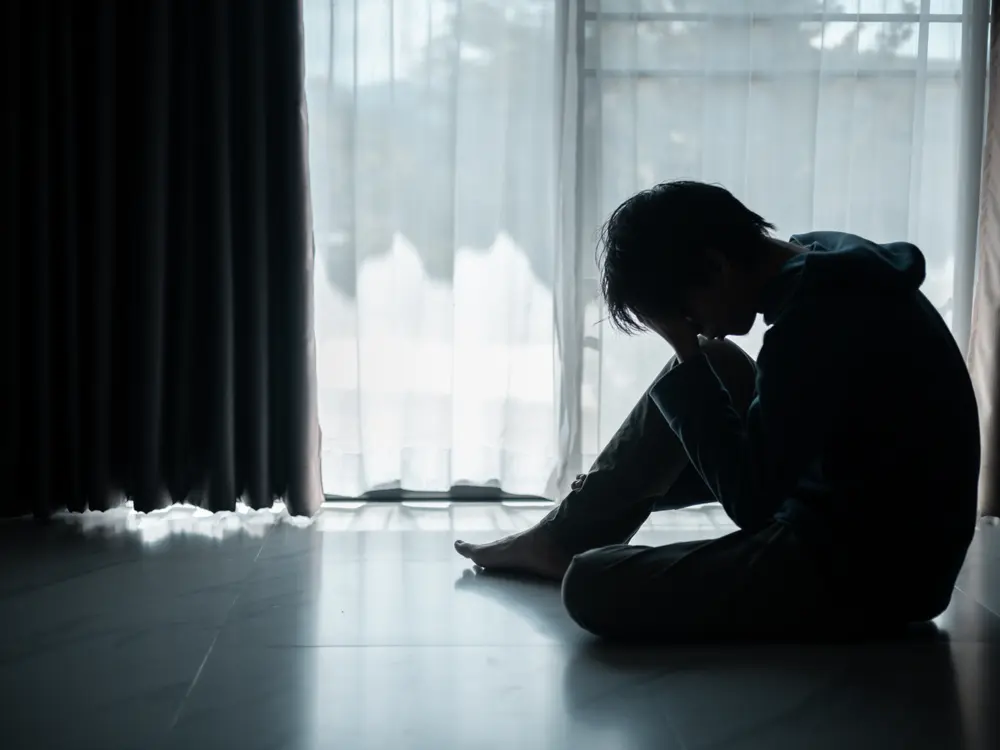 A young person sitting alone on the floor, head in hands, in front of a curtained window.